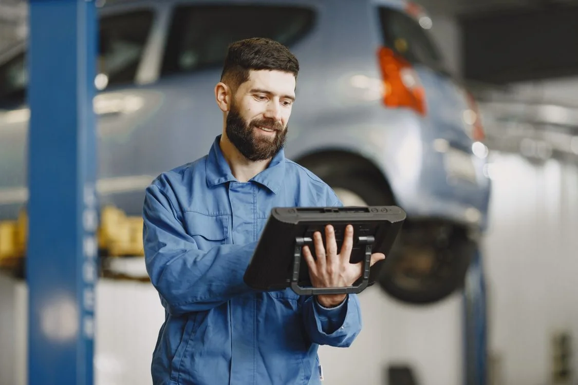 Mechanic in blue coveralls holding a tablet beside a vehicle on a lift